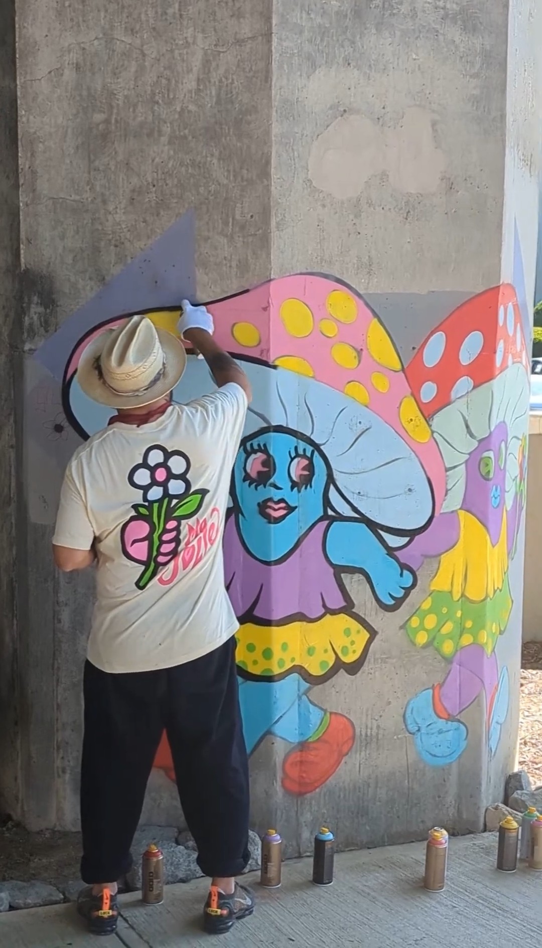 Artist painting a colorful character mural on a concrete pillar under a bridge.