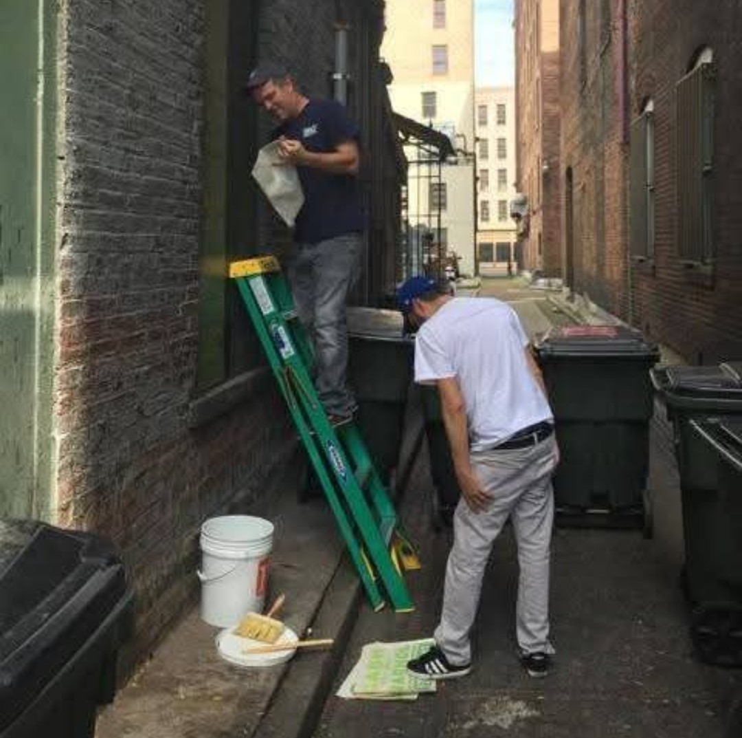 Two people preparing a wall for a mural in a Cincinnati alley, with a ladder, buckets, and prep materials.