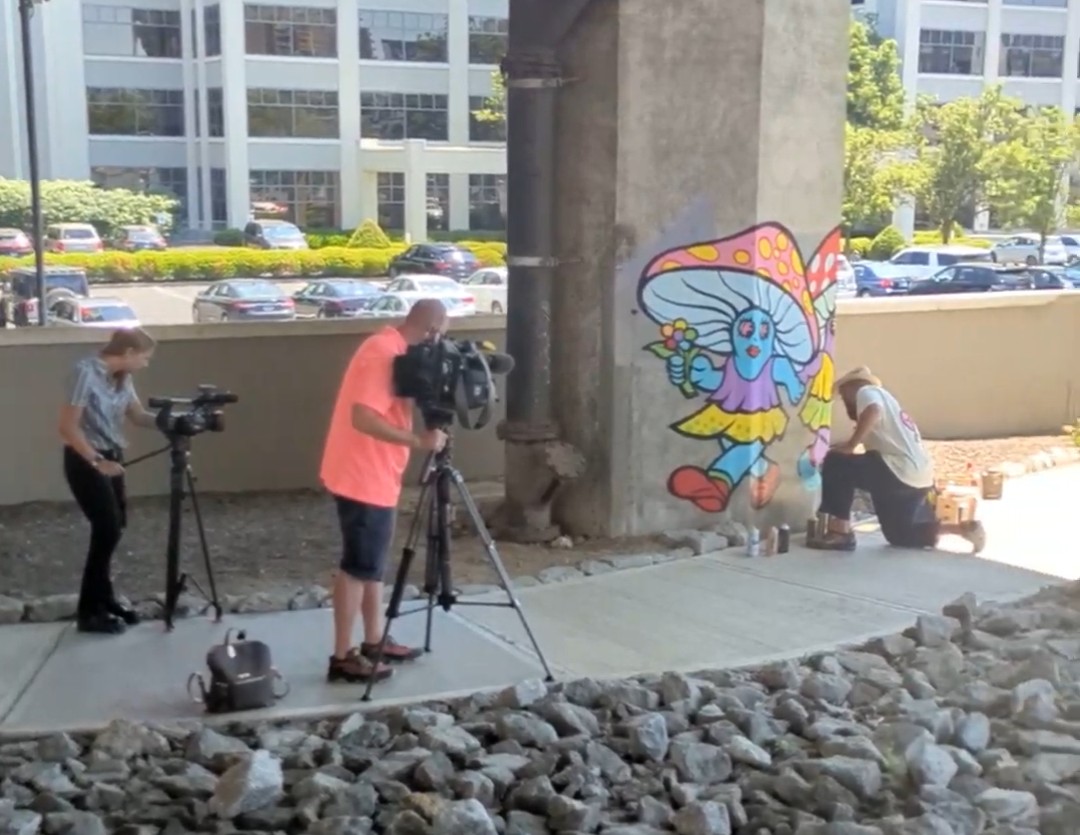 A camera crew filming an artist finishing a mural on a concrete pillar.